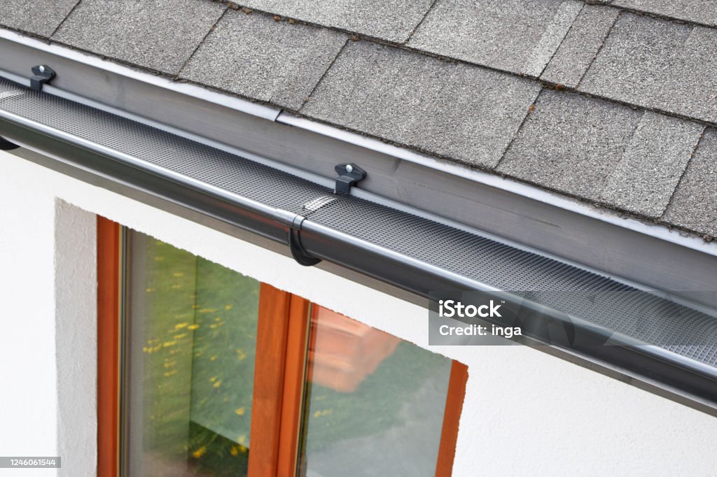 Plastic guard over new dark grey plastic rain gutter Plastic guard over new dark grey plastic rain gutter on asphalt shingles roof and white color decorative plaster facade with brown wooden window with reflection on glass pane. Roof Gutter Stock Photo