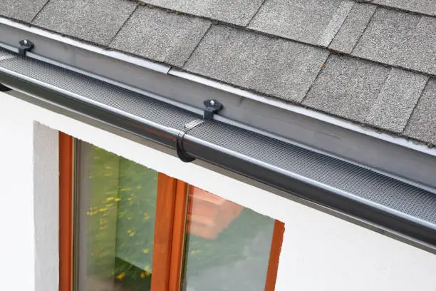 Plastic guard over new dark grey plastic rain gutter on asphalt shingles roof and white color decorative plaster facade with brown wooden window with reflection on glass pane.