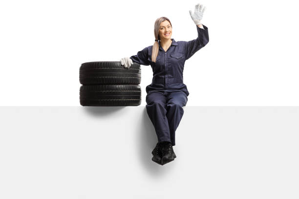 istock Female auto mechanic worker in a uniform sitting on a panel with tires and waving 1246090816
