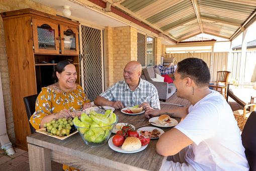 Young Pacific Islander couple and his mature father sitting outdoors eating together at the table. The young woman is pregnant.