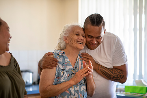 Senior Pacific Islander woman and her mature daughter and grandson cuddling each other in their kitchen at home.