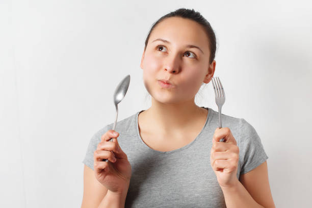 istock Girl with a thoughtful expression holding a fork and spoon. On a uniform light gray background 1246148187