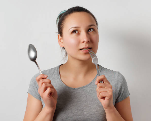 istock Girl with a thoughtful expression holding a fork and spoon. On a uniform light gray background 1246148193