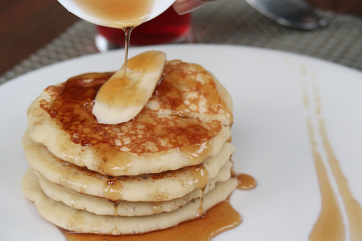 Stock photo showing some fresh scotch pancakes on a white plate, served with slices of banana and a generous amount of maple syrup being poured on top.