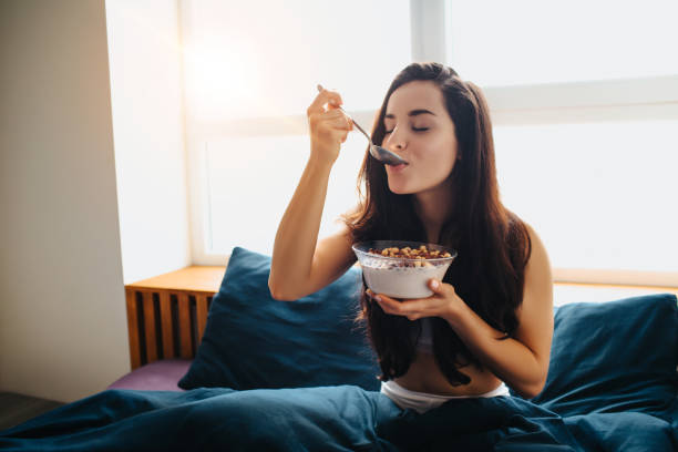 giovane bella donna a letto mattutino a casa. mangiare colazione ciotola di cereali al cioccolato con latte. gustando il gustoso cibo delizioso per la colazione del mattino. - eating women breakfast cereal foto e immagini stock