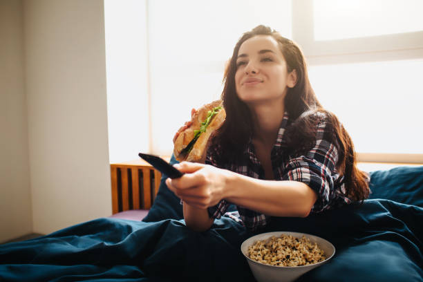 jovem linda mulher na cama matinal em casa. mulher feliz e animada assistindo tv. usando controle remoto e comendo sanduíche com pipoca. hora do filme. - freedom sandwich bread food - fotografias e filmes do acervo