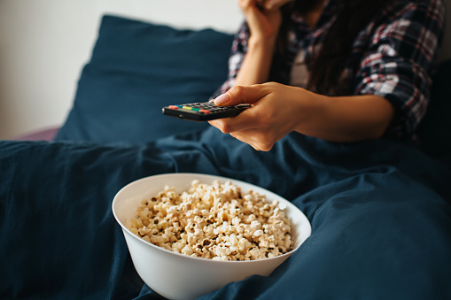 Young beautiful woman in morning bed at home. Hold remote control and watch tv or movie. Bowl with popcorn on bed. Cut view and close up.