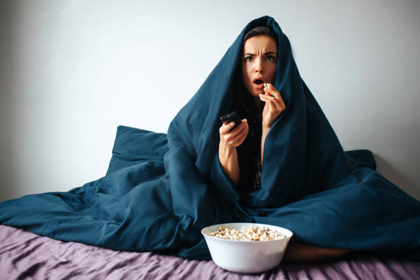 Young beautiful woman in morning bed at home. Watching scary movie alone in room. Covered with blanket. Eating popcorn and switching channels with remote control. Young beautiful woman in morning bed at home. Watching scary movie alone in room. Covered with blanket. Eating popcorn and switching channels with remote control part of a series stock pictures, royalty-free photos & images