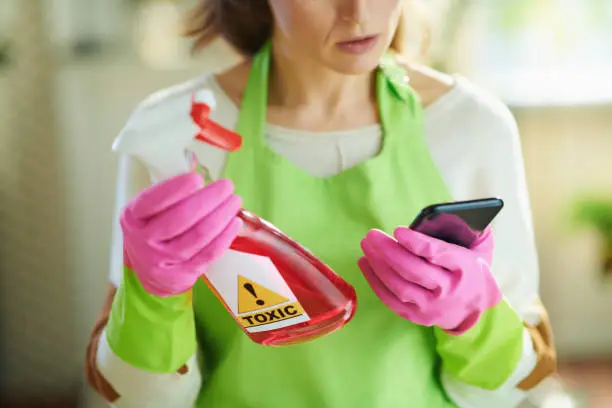 Closeup on woman in green apron and pink rubber gloves in the living room in sunny day reading about cleaning product on Internet using smartphone.