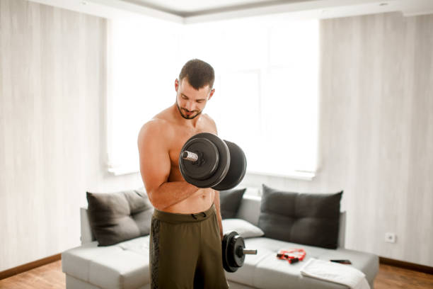 Young ordinary man go in for sport at home. View of hardwoking t-shirtless guy sportsman stand on floor and hold dumbbells in hand. Ready to start workout. Ordinary man work on improving his body el joven común común y corriente va a hacer deporte en casa. vista de los deportistas sin camiseta de mujer se paran en el suelo y sostienen las mancuernas en la mano. listo para empezar el entrenamiento. el hombre ordinario trabaja en mejorar su cuerpo.