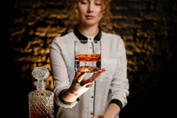 giovane bella barista tiene un bicchiere vecchio stile con bevanda alcolica - gin decanter whisky bottle foto e immagini stock