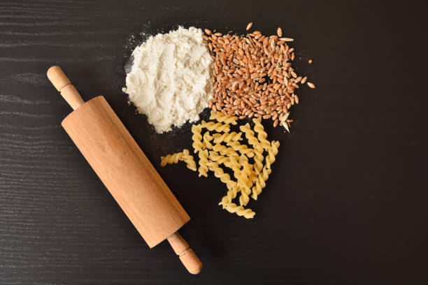 White flour, wheat grains, raw macaroni or pasta, and rolling pin on wooden black desk with copy space for design or text. Domestic food concept. Close up, selective focus and top view