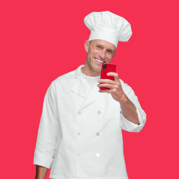 istock Caucasian young male chef standing in front of colored background wearing jacket and using mobile phone 1246294446