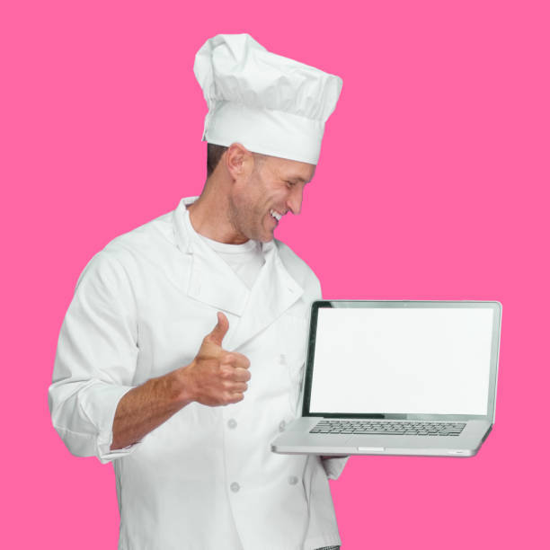 istock Caucasian male chef standing in front of colored background wearing uniform and using laptop 1246294828
