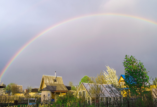 Rainbow over the roofs of rural houses in the stormy evening sky after a storm, summer village landscape.