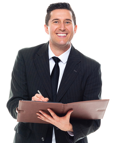 One person of caucasian male businessman standing in front of white background wearing businesswear who is cheerful who is writing and holding book and using pen