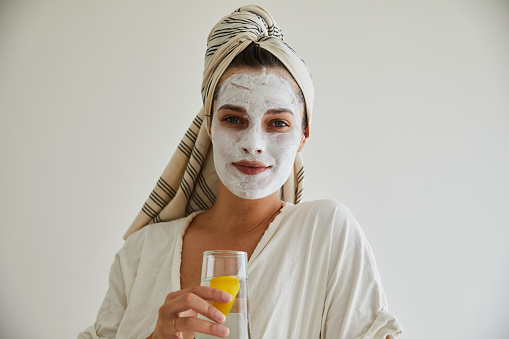 Portrait of a smiling young woman in a bathrobe and wearing a cleansing face mask drinking a glass of water