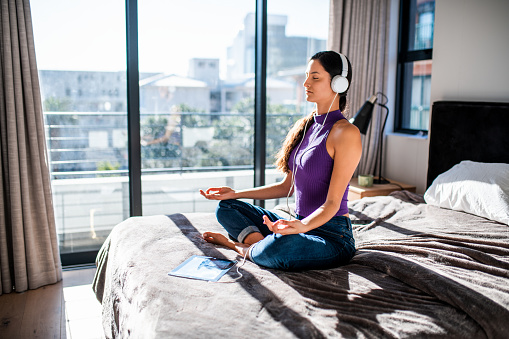 Young Latin woman meditating in the bed on a beautiful sunny morning. She is wearing headphones.