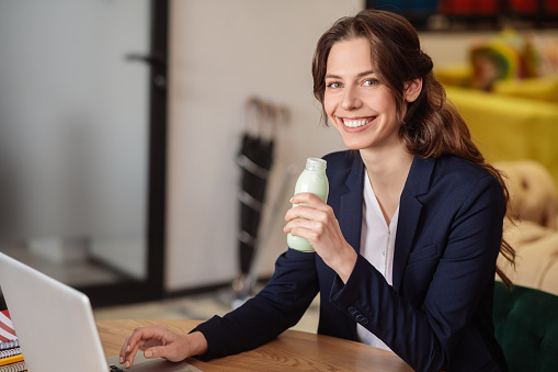 Healthy drink. Smiling long-haired girl with a small bottle of yogurt in her hand sitting at a laptop in the office