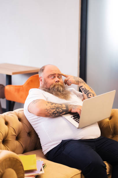 istock Bald bearded plump man sitting with a laptop in hands and looking thoughtful 1246396526