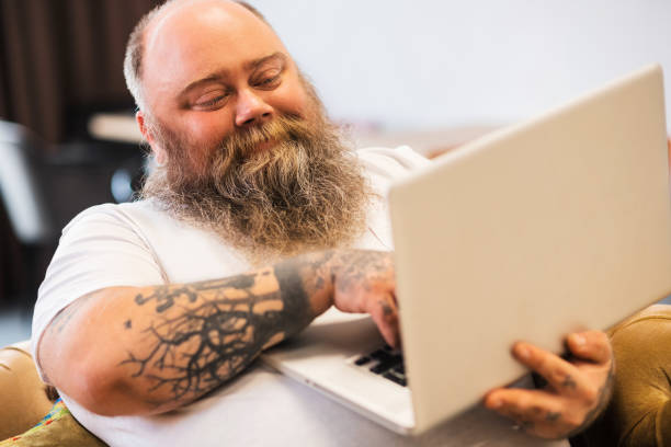 istock Bald bearded plump man sitting with a laptop in hands and smiling 1246396527