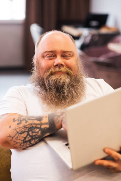 istock Bald bearded plump man sitting with a laptop in hands 1246396528