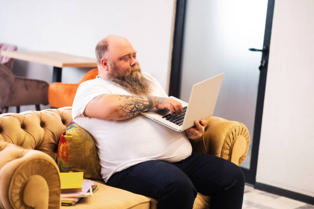 istock Bald bearded plump man sitting with a laptop in hands and working online 1246396530