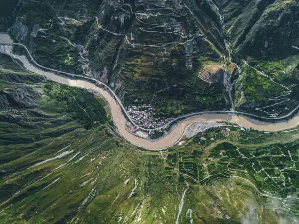 Top down aerial drone view of river and small settlement in between the tall mountains in Tibetan area of Sichuan province, China. Cinematic shot, curved serpentine roads and hills. Journey in spring.