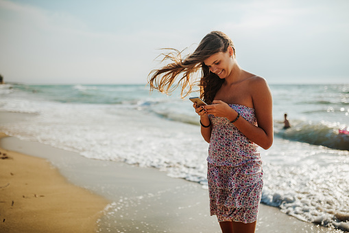 Beautiful young woman in dress standing on the beach and texting message on smart phone. Summer vacation and social media concept