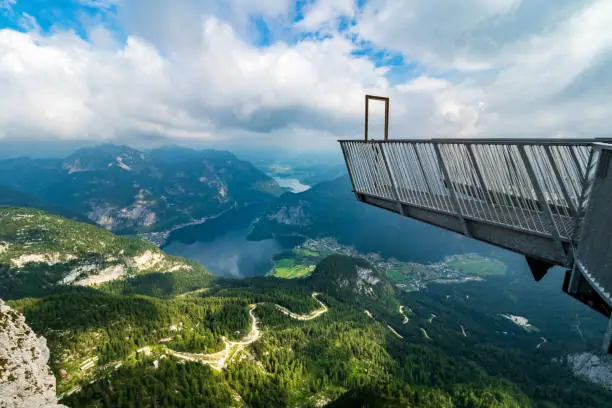 Krippenstein, Austria - July 16th 2019: From the 5 Fingers, a steel construction on top of the Krippenstein Mountain at the Dachstein glacier massif, tourists and hikers can enjoy an unique and amazing view of the Salzkammergut region with it's beautiful lakes and mountains. Clouds at the same altitude as this observation point give the picture a dramatic note. Down at the valley, the Lake Hallstatt is visible, with the famous mountain village Hallstatt at it's shore. 
At the end of this platform, a big painting frame is located, allowing people to take beathtaking selfies.