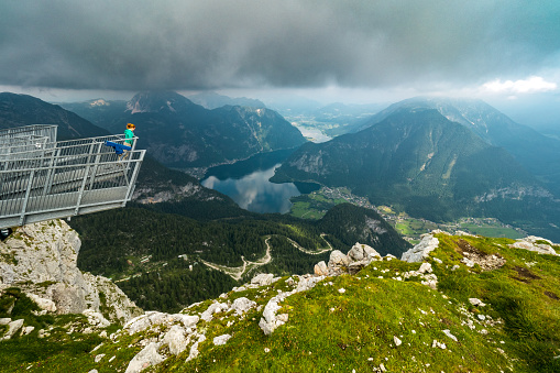 Krippenstein, Austria - July 16th 2019: From the 5 Fingers, a steel construction on top of the Krippenstein Mountain at the Dachstein glacier massif, tourists and hikers can enjoy an unique and amazing view of the Salzkammergut region with it's beautiful lakes and mountains. Clouds at the same altitude as this observation point give the picture a dramatic note. Down at the valley, the Lake Hallstatt is visible, with the famous mountain village Hallstatt at it's shore. \nA woman stands alone at the platform and enjoys the thrilling view.