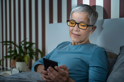 Senior woman with blue-light blocking glasses lying in bed before sleeping, using smartphone.