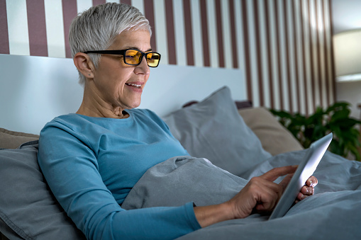 Mature woman wearing blue light blocking glasses with amber lenses, lying in bed before sleep, looking at tablet screen