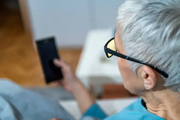 Using Blue-Light Blocking Glasses in the Evening for Better Sleep. Mature Woman lying in bed, holding smart phone