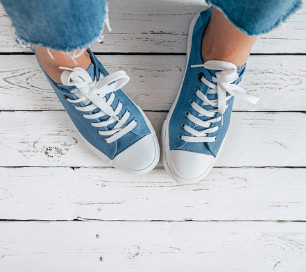 Teenager's feet in blue color casual new sneakers with white shoelaces on the white wooden floor close up image. Vintage style in modern fashion world concept image.