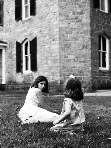 Two sisters sitting on the grass and playing next to the old stone house, eldest sister looking at camera, youngest- 
she sits with her back to us. Black and white photo.