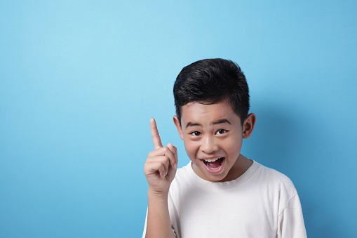Portrait of happy cheerful Asian boy pointing finger up, having idea gesture with big smile, against blue background