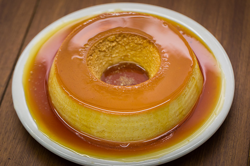 Pudim de leite, a delicious Brazilian flan dessert, with milk ,eggs and condensed milk, topped with caramel sauce.( It's type of vanilla pudding). Served on white plate on wooden table.