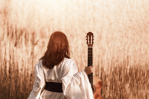 Beautiful young woman with red hair in a white medieval dress holding guitar and walking through the sunny field at warm light of sunset. Lens flare, toned image, selective focus, blurred bokeh.