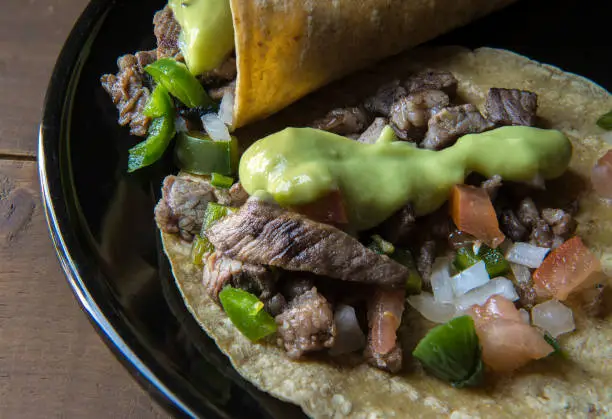 Mexican beef tacos, or Mexican taquitos with salsa and avocado cream or guacamole on a black plate on a wooden table, tacos made with corn tortillas, spicy pico de gallo or salsa Bandera