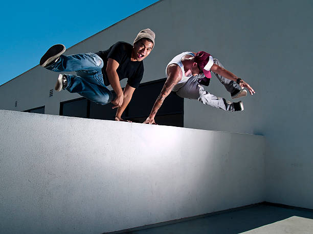 Two Male Freerunners Jumping Over a Wall Performing Parkour Two young male parkour freerunners jumping over a wall. free running stock pictures, royalty-free photos & images