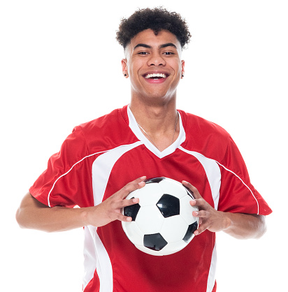 Waist up of aged 18-19 years old with black hair african ethnicity male athlete standing in front of white background wearing soccer uniform who is showing cool attitude and holding soccer ball and playing soccer - sport and using sports ball