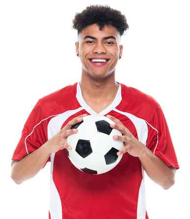 Portrait of aged 18-19 years old with curly hair generation z young male athlete standing in front of white background wearing soccer uniform who is happy and holding soccer ball and playing soccer - sport and using sports ball