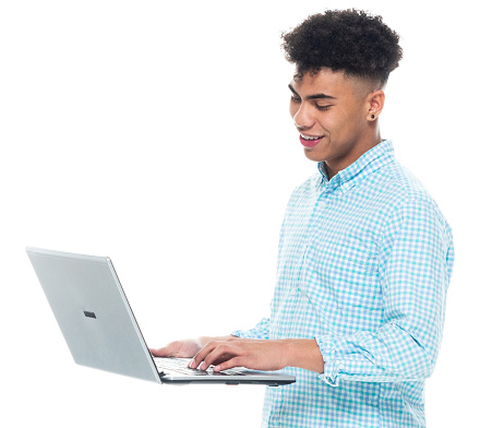 Side view of aged 18-19 years old with black hair african ethnicity young male standing in front of white background wearing rolled-up sleeves who is learning who is working and using laptop