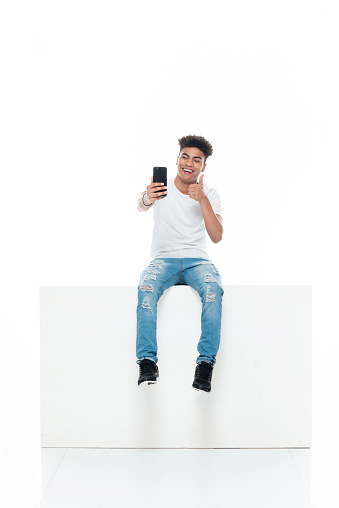 Full length of aged 18-19 years old with curly hair generation z teenage boys photography resting in front of white background wearing shirt who is showing cool attitude who is taking a selfie and using mobile phone