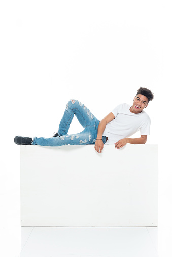 Profile view of aged 18-19 years old with curly hair african ethnicity teenage boys lying down in front of white background wearing t-shirt who is laughing