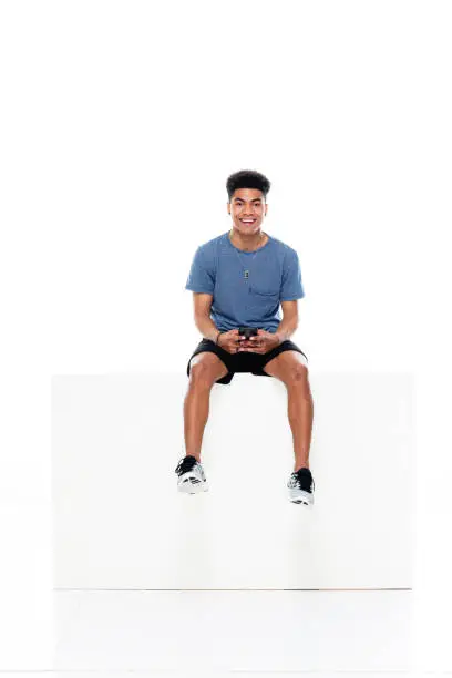 Full length of aged 18-19 years old with black hair african ethnicity male sitting in front of white background wearing t-shirt who is showing cool attitude and holding box and using mobile phone