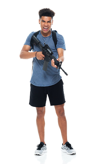 One person of aged 18-19 years old with black hair generation z young male criminal standing in front of white background wearing shorts who is angry who is violence and holding weapon and using rifle