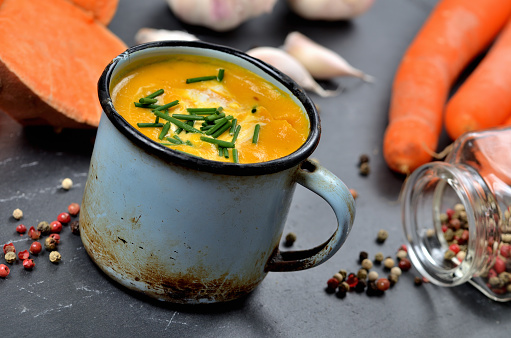 Sweet potato soup with cream and fresh chive in old enamel cup. Garlic, pumpkin, spilled colored pepper and half pieced carrot in background on slate slab.