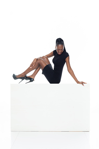 Profile view of aged 20-29 years old who is beautiful with long hair african-american ethnicity young women resting in front of white background wearing dress and high heels who is happy and holding box